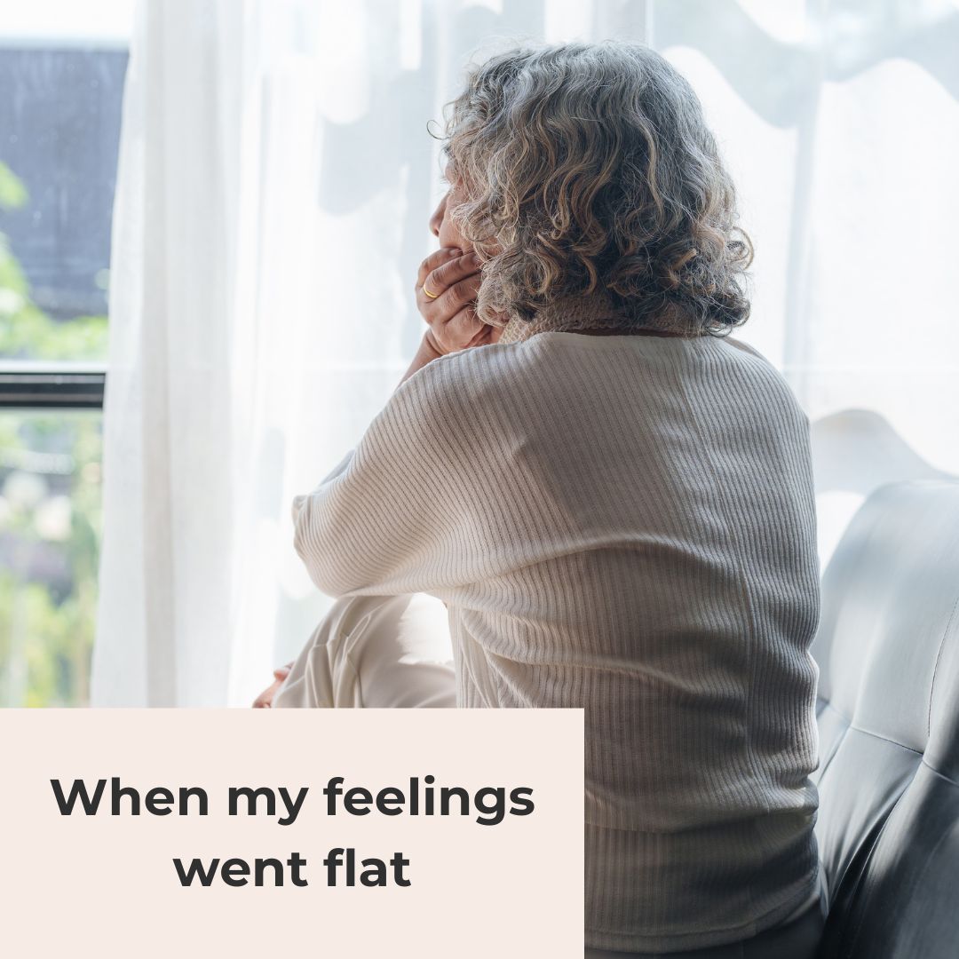 A woman with curly silver hair sits curled up in a chair by a window, looking out through a sheer curtain, in a post about low mood in perimenopause.